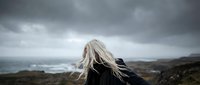 Young blonde woman with flowing hair standing on dramatic coastal cliff overlooking stormy ocean waves under moody overcast sky for travel and adventure concepts.