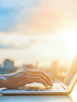 Male hands typing on laptop keyboard at sunset with urban cityscape background and warm sunlight streaming through window, creating atmospheric workspace scene.