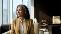 Middle-aged woman with long dark hair and East Asian ethnicity, wearing a tan blazer and white shirt, sitting in a modern high-rise office with city skyline views, professional business environment.