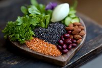 Heart-shaped wooden plate with healthy ingredients including black caviar, red lentils, almonds, fresh herbs, and kidney beans arranged artistically for food styling.
