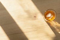 Glass of whiskey with ice cube on wooden table casting dramatic diagonal shadows in sunlight, creating minimalist composition with warm tones.