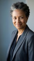 Professional African American businesswoman with gray curly hair smiling confidently in dark suit jacket against neutral background for corporate headshot.