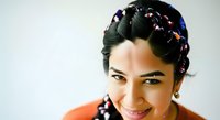 Young woman with dark hair styled in braids with colorful beads, wearing orange top, smiling at camera against white background. Portrait for beauty, lifestyle, and diversity content.