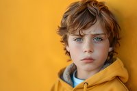 Young boy with striking blue eyes wearing yellow hoodie against vibrant yellow background, close up portrait showing innocence and natural expression.