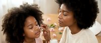 Young African American mother applying makeup to daughter's face during beauty bonding time at home, teaching skincare and cosmetics application for special occasions.