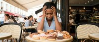 Stressed young African American woman holding head in hands at cafe table with pastries and coffee, expressing overwhelm and fatigue in restaurant setting.