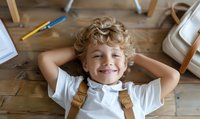 Young caucasian boy with curly blonde hair and bright smile wearing white t-shirt and backpack lying on wooden floor during study break at home, education concept.