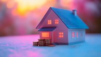 Miniature house model with glowing windows and stacked coins against pink sunset background, representing real estate investment and home financing concept.