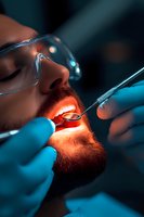 Caucasian male patient with red beard receiving dental treatment while wearing protective glasses. Dentist hands in blue gloves holding dental tools in dark setting.