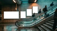 Modern subway station interior with escalators and blank illuminated advertising billboards for commercial messaging and branding opportunities.
