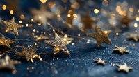 Golden star confetti scattered on dark blue glittery surface with bokeh lights in background, creating festive holiday atmosphere.