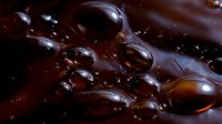 Dark brown liquid surface with glossy bubbles and droplets creating abstract macro photography pattern with rich chocolate or coffee tones and reflective highlights.