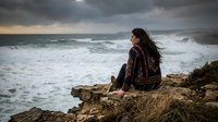 Young woman with long dark hair sitting on rocky cliff overlooking stormy ocean waves under dramatic cloudy sky during golden hour sunset.