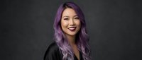 Young Asian woman with vibrant purple hair smiling confidently in black clothing against dark background for fashion and beauty concepts.