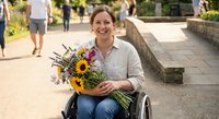 Smiling woman in wheelchair holding colorful sunflower bouquet outdoors in urban park setting with stone pathways and greenery in background.