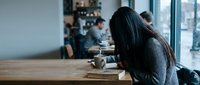 Young Asian woman in gray sweater sitting at wooden table with coffee cup in modern cafe, reading book during daytime with natural window light and blurred background.