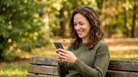 Happy young woman with curly brown hair using smartphone while sitting on wooden bench in sunny autumn park with golden foliage background.