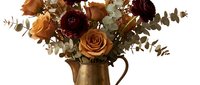 Autumn floral arrangement with orange and burgundy roses in vintage brass pitcher with eucalyptus leaves on white background for seasonal decor.