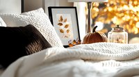 Cozy autumn home decor with framed dried leaves artwork, decorative pumpkin and glass jar on white bedding, creating warm fall atmosphere with golden bokeh light.