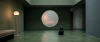 Young person sitting alone in minimalist gallery space with glowing circular light installation on dark wall, contemporary art exhibition interior design.