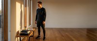 Young Caucasian man in black casual outfit standing in modern minimalist room with wooden floor and large windows creating warm natural lighting.
