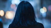 Young Asian woman with dark hair from behind against blurred city bokeh lights background creating moody atmospheric evening scene.