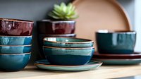 Handcrafted ceramic bowls and plates in teal blue and burgundy glazes stacked on wooden surface, with small succulent plant decoration. Modern pottery dinnerware set.