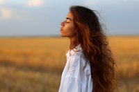 Young woman with flowing long brown hair in white shirt standing in golden wheat field at sunset, eyes closed in peaceful meditation moment.