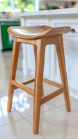 Modern wooden bar stool with curved seat in natural bamboo finish, photographed in bright kitchen setting against white cabinets and tile floor.