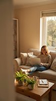 Young Caucasian woman working on laptop computer while sitting comfortably on beige sofa in bright modern living room with natural sunlight streaming through window.