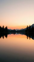 Serene mountain lake at sunrise with golden sky reflecting on calm water, surrounded by evergreen forest silhouettes and distant peaks for nature and travel projects.