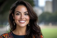 Young woman with long dark wavy hair and warm smile wearing colorful patterned jacket outdoors in park setting with blurred green background, suitable for lifestyle and portrait use.