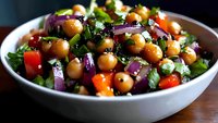 Fresh chickpea salad with colorful bell peppers, red onions, and herbs in white ceramic bowl, garnished with chopped parsley and olive oil dressing.