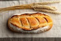 Fresh baked artisan sourdough bread loaf with golden brown crust and scoring pattern on rustic linen cloth background with wheat stalks, top view.