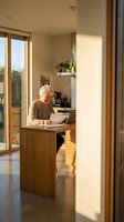 Senior Caucasian woman working on laptop at wooden kitchen island in modern home with warm natural lighting and large windows during golden hour.