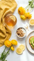 Bright flat lay breakfast scene with lemon tea, granola yogurt bowl, avocado toast, and yellow ranunculus flowers on marble.