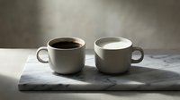 Two ceramic mugs on marble surface, one filled with black coffee and one empty, morning beverage and kitchenware styling for cafes and home decor.