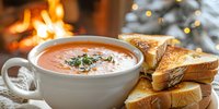 Warm tomato soup in white bowl served with toasted bread slices, fireplace glow and decorated tree in background, comfort food for winter holidays.