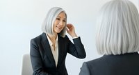Mature woman with gray hair wearing black blazer and white shirt, touching face while looking at mirror reflection against white background.