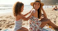 Young woman applying sunscreen on friend's back at beach during golden hour vacation moment with ocean waves and sandy shore.