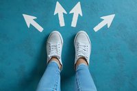 White arrows pointing in different directions around casual sneakers on turquoise background. Person wearing light blue jeans standing at crossroads making choice.