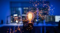 Glowing light bulb with sparkling electrical energy bursts against blue office background, representing innovation, creativity, and bright ideas for business concepts.