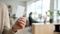 Close-up of hand holding blank white card in modern office environment with colleagues working at desk in background, suitable for business branding and corporate messaging.