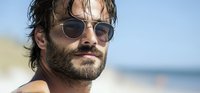 Bearded man wearing sunglasses at beach on sunny summer day, with wet hair and confident expression, looking away from camera against blurred ocean background.