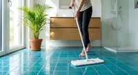 Woman mopping turquoise tile bathroom floor with white mop near modern vanity and palm plant. Household cleaning and maintenance routine.