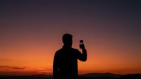 Man silhouette taking selfie photo with smartphone against dramatic sunset sky with orange and purple gradient colors for social media content.