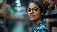 Young Hispanic woman wearing glasses and headband smiling in warehouse environment with blurred storage shelves background, casual plaid shirt, natural makeup look.