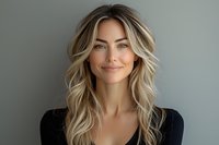 Young Caucasian woman with wavy blonde hair and natural makeup smiling confidently against gray background, wearing black top. Professional headshot style portrait.