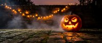 Glowing jack-o-lantern with carved face sits in misty autumn landscape at dusk, surrounded by warm string lights and dark trees creating spooky seasonal atmosphere.