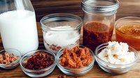 Variety of homemade fermented foods in glass jars on wooden table with milk bottle, including kimchi, sauerkraut and pickled vegetables for gut health.